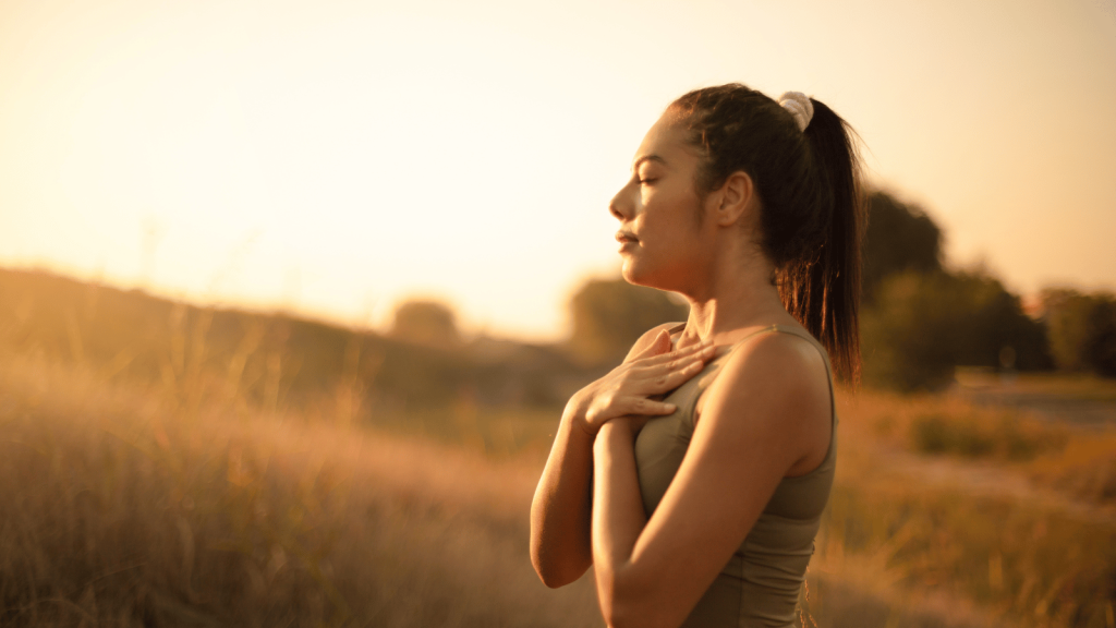 A woman outdoors during golden hour stands with her eyes closed and her hands resting gently on her chest. She appears calm and peaceful in a field with warm sunlight behind her.