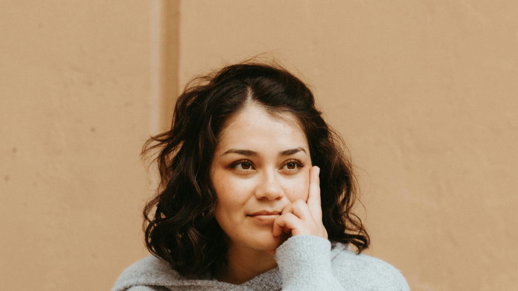 A woman with shoulder-length dark wavy hair rests her face on her hand while looking off thoughtfully. She wears a soft light-gray sweater and sits against a plain tan background.