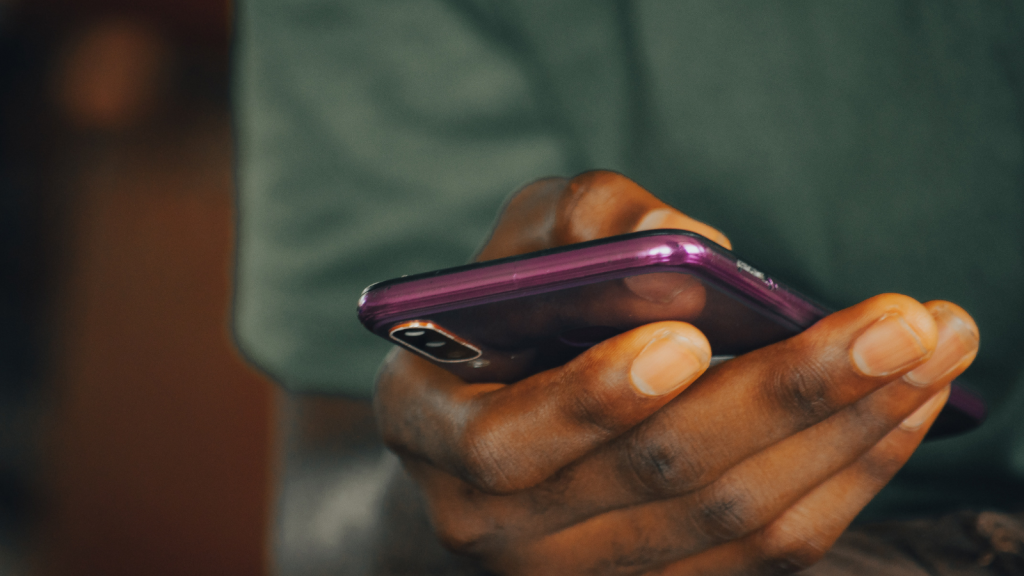 A close-up of a person holding a purple smartphone in one hand, with their thumb positioned near the edge of the device. The background is dark and blurred.