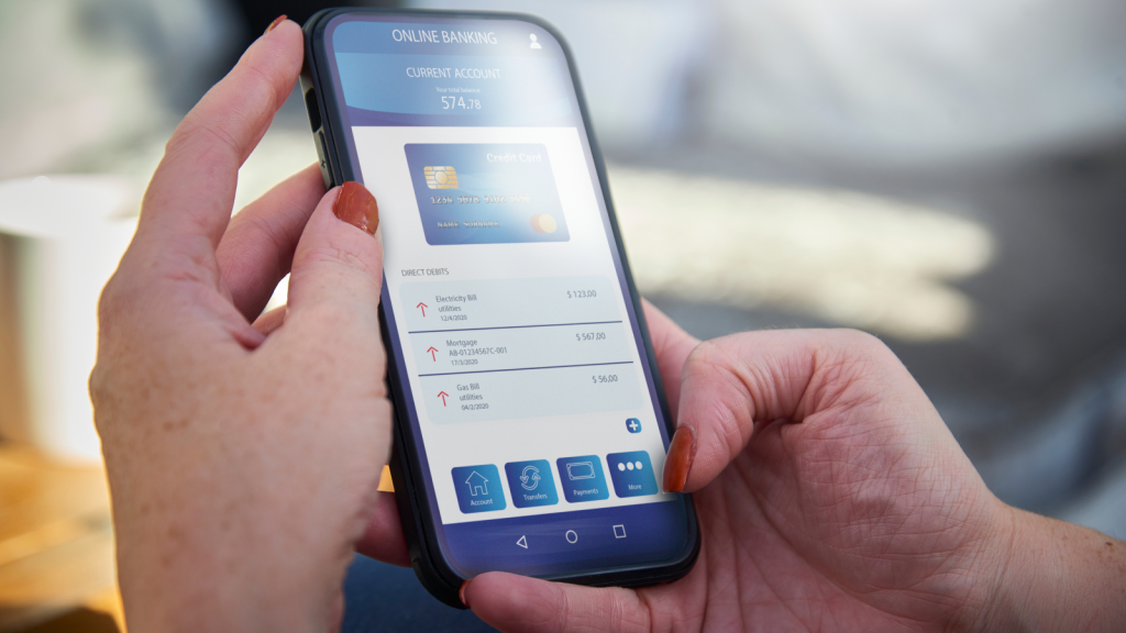 A person holding a smartphone displaying an online banking app with a balance of $574.78 and several direct debit entries. The phone is held with both hands, and the app interface shows icons for account, transfers, payments, and more. The person has light skin and brown nail polish.