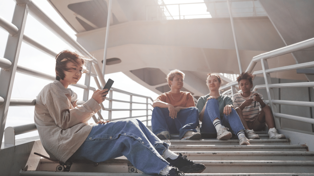 Four teenagers lounge on outdoor metal stairs. One sits separately, looking at a phone, while the other three sit together talking. Sunlight streams through the railings.
