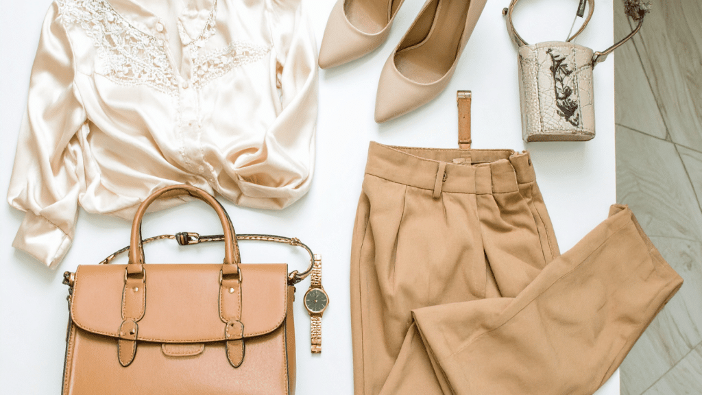 A flat lay of coordinated beige and tan fashion items: a silky cream blouse with lace detailing, tan trousers, beige pointed-toe heels, a structured tan handbag, a gold wristwatch, and a small silver textured handbag with a floral design. Arranged neatly on a white surface.