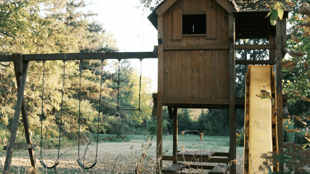 An old wooden backyard playset with swings, rings, and a yellow slide stands in a grassy yard surrounded by trees. The structure looks weathered and unused.