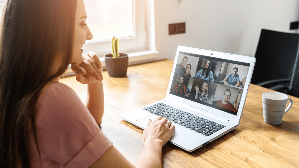 A woman sits at a table using a laptop for a multi-person video call. Several faces appear on the screen, and a mug and small plant sit nearby. The room is softly lit by daylight.