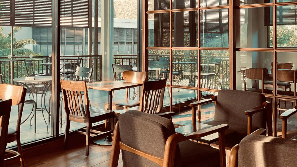 A quiet café interior with wooden tables and chairs arranged near large windows. The afternoon sunlight creates warm shadows across the floor and furniture.