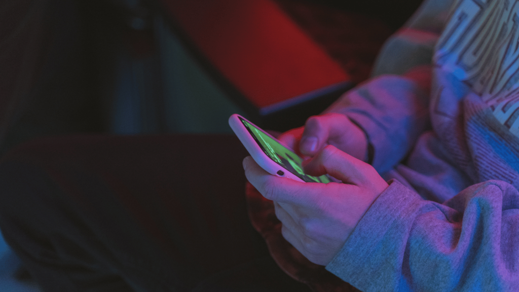 A person in a hoodie holds a smartphone with both hands in a dimly lit room, illuminated by green and blue light from the screen.