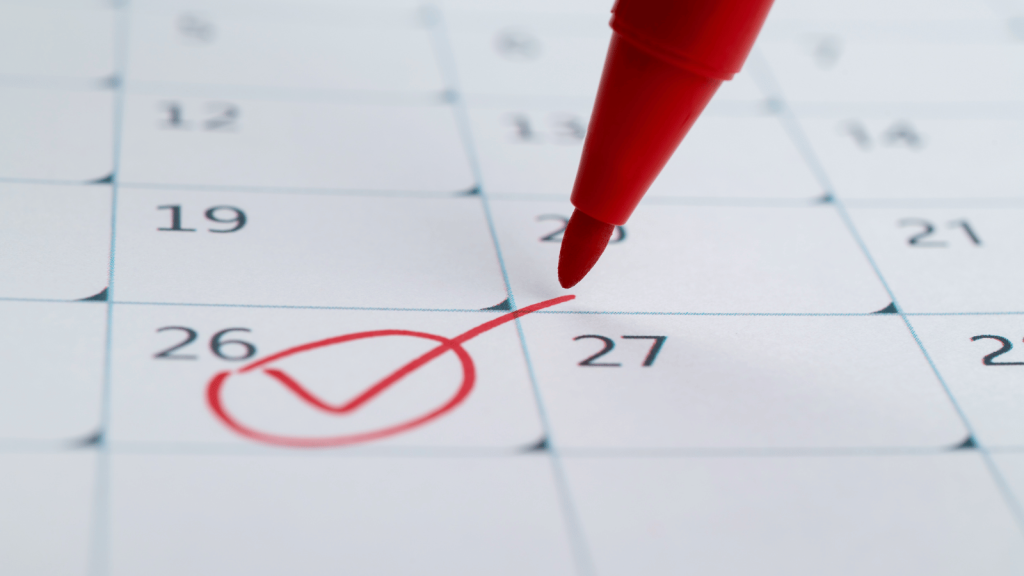 A calendar page with a red checkmark drawn over the 26th. A red marker touches the paper near the marked date.
