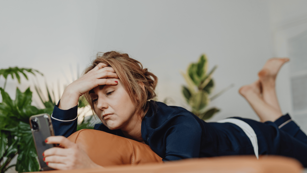 A woman in navy pajamas lies on her stomach on a couch, holding her phone and resting her hand on her forehead in fatigue. Green houseplants fill the background.