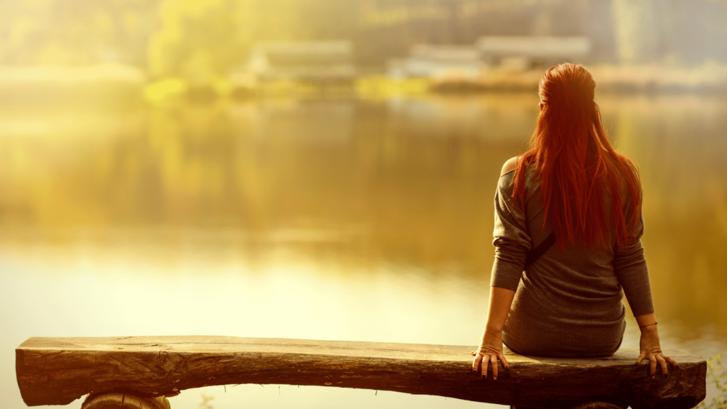 A woman with long red hair sits alone on a wooden bench, facing a calm lake in warm golden light.