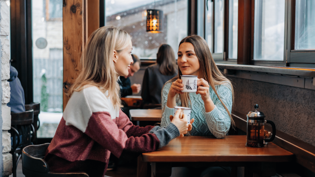 Two women sit across from each other in a cozy café, smiling and talking while holding mugs. Snow is visible outside the window, and other patrons sit in the background.