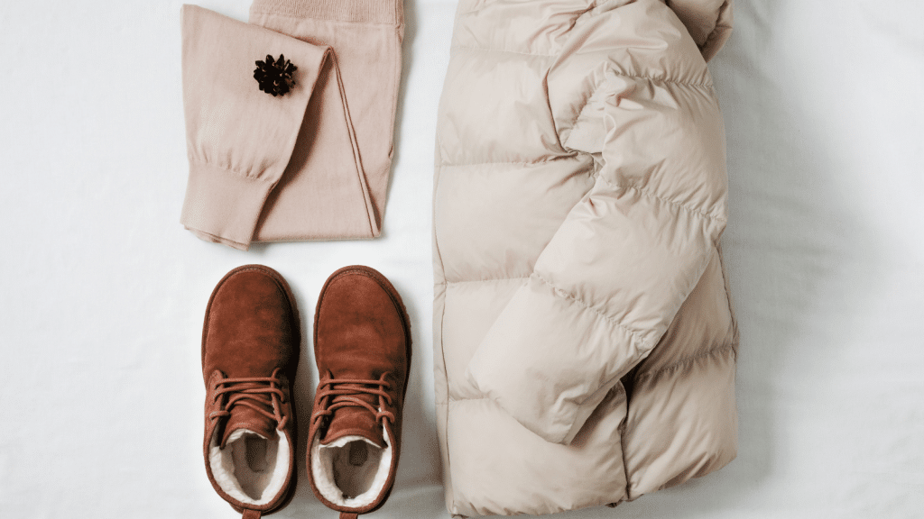 A flat lay of neutral winter clothing, including folded beige knit pants with a dark brown pinecone accessory, a cream-colored puffer jacket, and a pair of brown suede lace-up boots lined with light fleece. All items are arranged neatly on a white surface.
