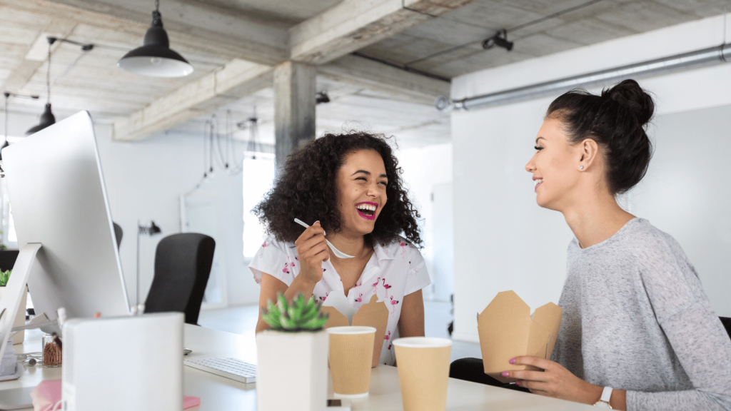 Two women sit together at an office desk, laughing while eating takeout food. One holds a spoon and the other holds a food container. Coffee cups, a plant, and a computer monitor are on the table.