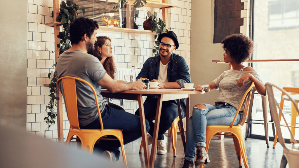 Four people gather around a round table in a bright café, engaged in lively conversation. They sit on yellow chairs, and plants decorate the tiled wall behind them.