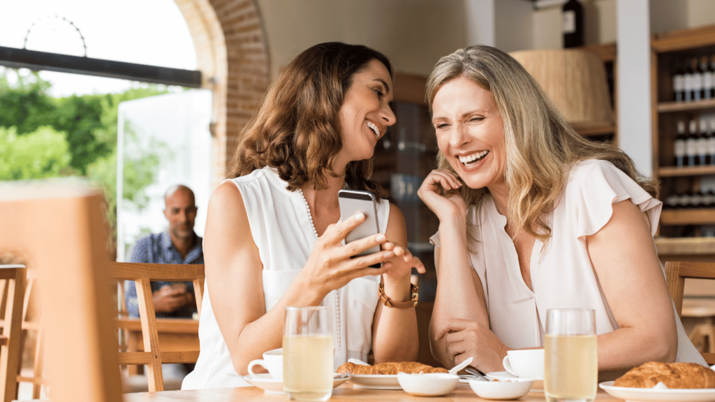Two women sitting at a café table, laughing together as one shows something on her phone; cups of juice and pastries are on the table.