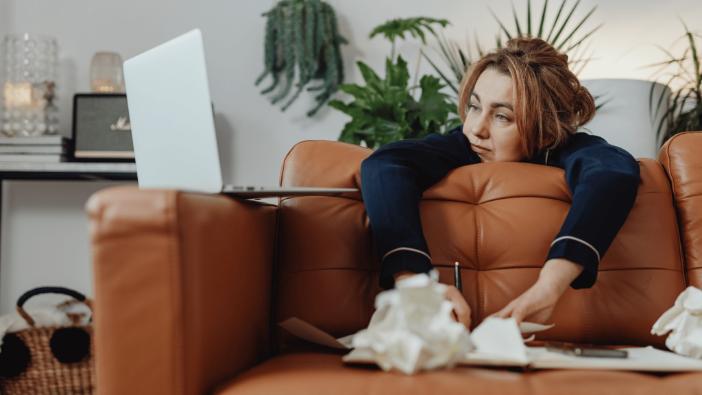 A woman in dark pajamas slumps over the back of a leather couch, staring tiredly at an open laptop. Crumpled tissues and scattered papers lie on the table in front of her, with plants and home décor in the background.
