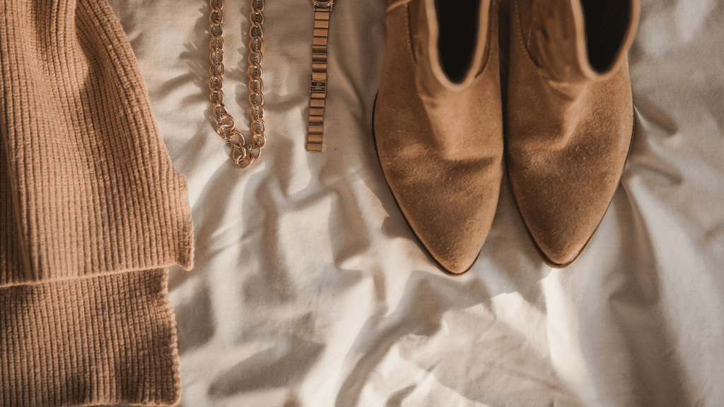 A soft, warm-toned flat lay featuring a ribbed beige knit garment on the left, a gold chain necklace and gold watch in the center, and a pair of tan suede pointed-toe ankle boots on the right. The items rest on a lightly wrinkled cream fabric.