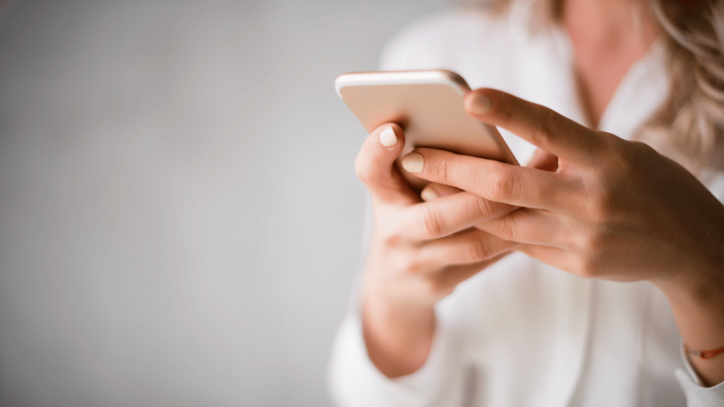 A close-up of a person holding a smartphone in both hands. The individual wears a white long-sleeve top, and the background is softly blurred. Only their hands and part of their torso are visible, focusing attention on the phone.