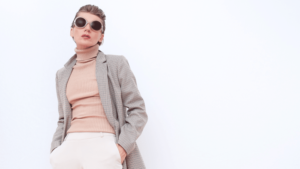A woman dressed in a beige ribbed turtleneck, cream trousers, and a long houndstooth blazer stands against a plain white background. She wears oversized round sunglasses and has her hands in her pockets, posing confidently while looking slightly upward.