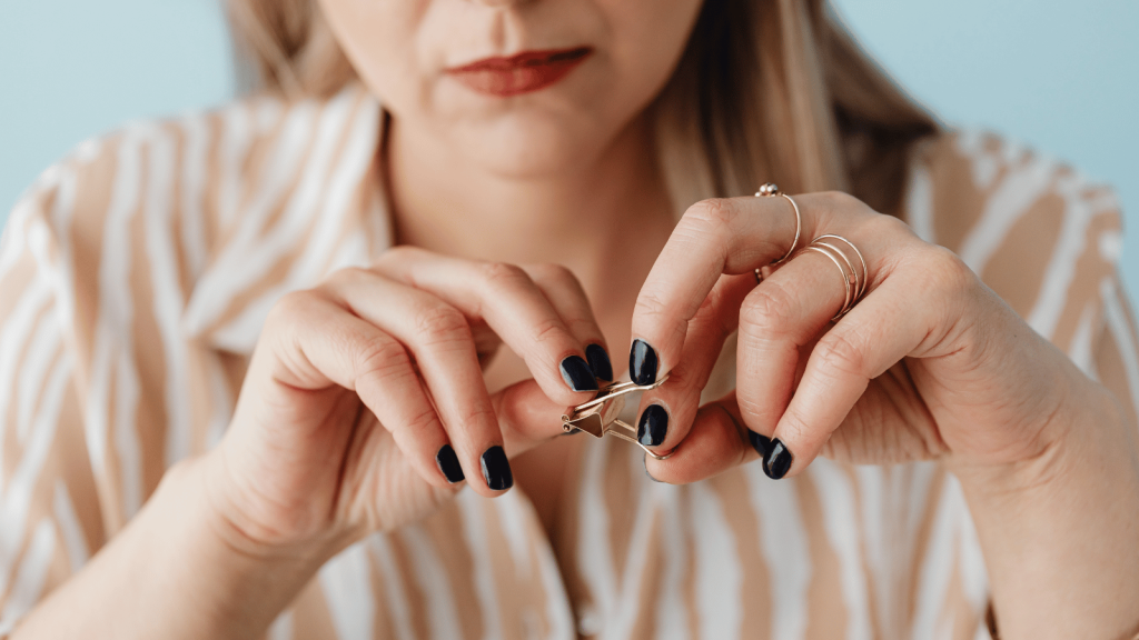 A woman fidgets with a pair of rings in her hands, her dark-painted nails and striped blouse visible, conveying quiet discomfort.