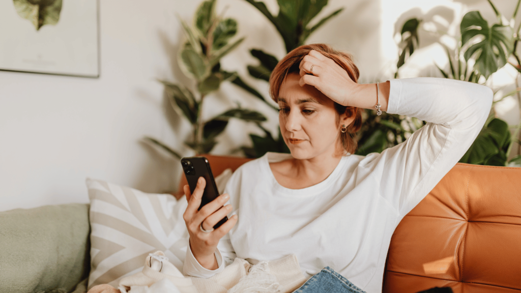 A woman sits on a sofa, leaning forward with her phone in hand, her other hand pressed to her forehead in concern. She is surrounded by plants and soft home décor.