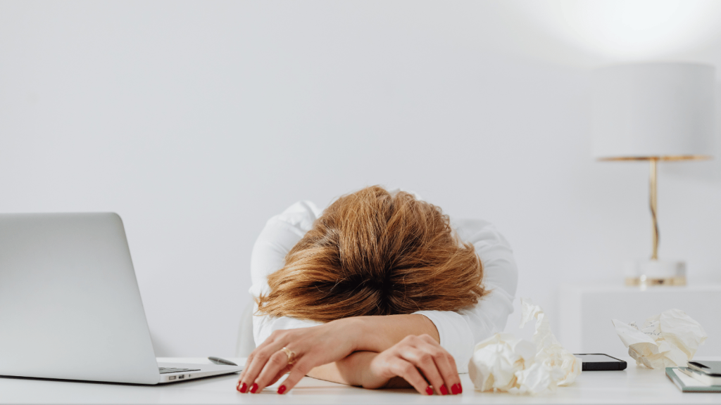 A person with their head down on a desk, arms folded under their forehead, sits near a laptop, a phone, and several crumpled tissues in a bright white workspace.