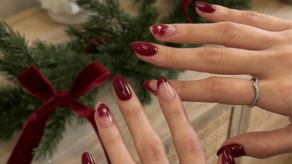 Glossy burgundy nails paired with nude accent nails featuring diffused burgundy floral-style art with tiny gold centers. Holiday greenery in the background reinforces the Christmas nails theme, while the refined palette fits old money Christmas nails.
