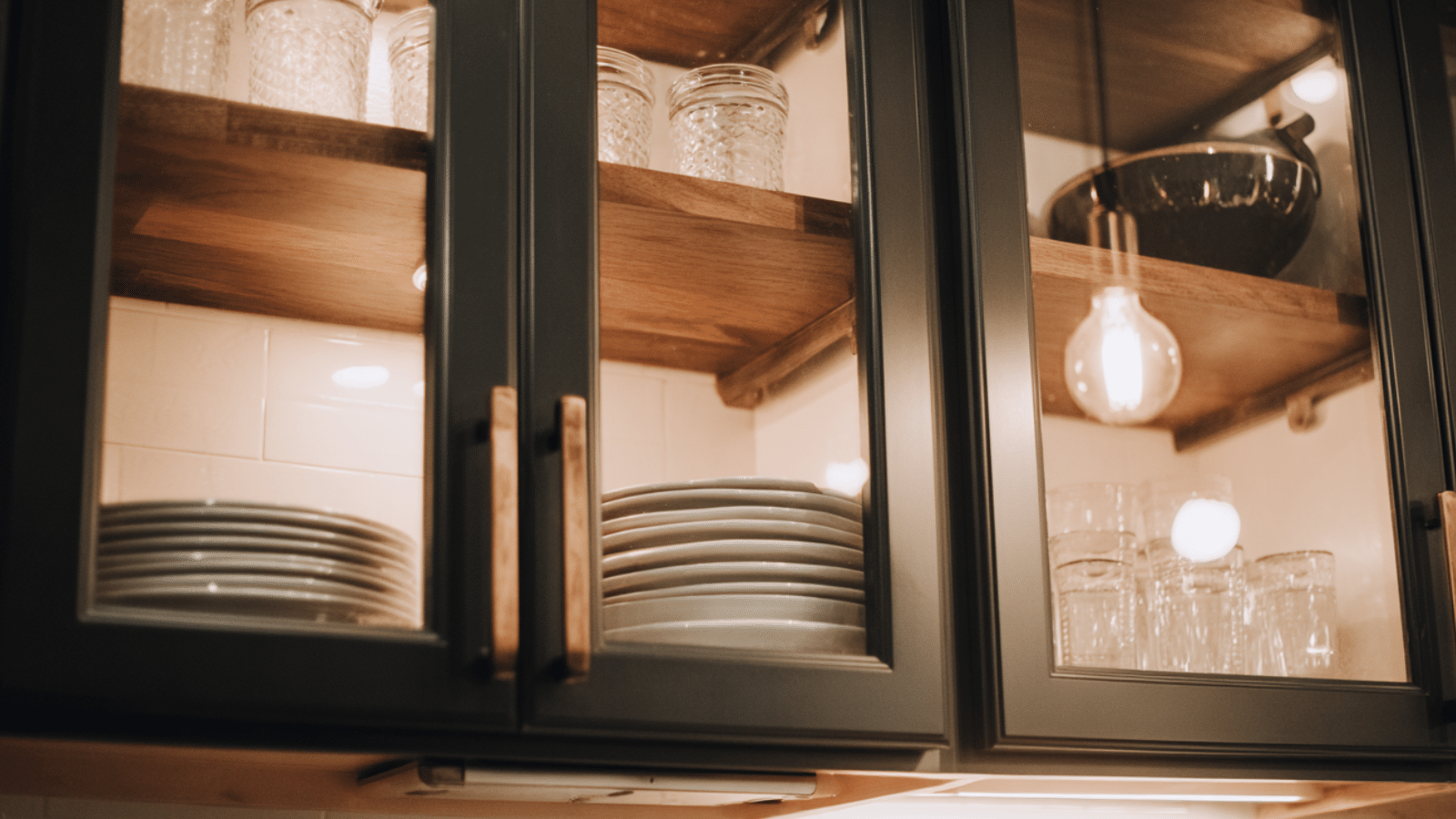 Glass-front kitchen cabinets with wooden shelves, neatly stacked white plates and clear drinking glasses, softly lit by warm interior cabinet lighting.